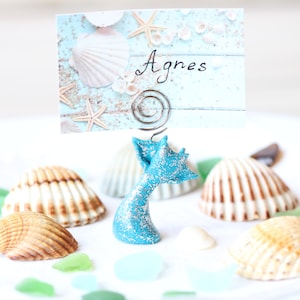 May include: A turquoise mermaid tail place card holder with a silver spring clip holding a card that says "Agnes" on a blue and white background with seashells and starfish. The holder is surrounded by seashells and sea glass on a white plate.