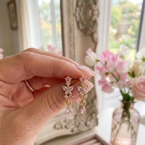 May include: A pair of gold-tone drop earrings featuring a butterfly motif. Each earring is adorned with small clear stones and a single pink teardrop-shaped stone at the base, suspended from a delicate gold-tone chain link.