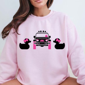 Peut inclure: Sweat-shirt rose avec une Jeep noire et deux canards en caoutchouc roses avec des nœuds sur la tête.