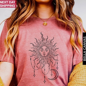 Könnte beinhalten: Ein kurzärmeliges T-Shirt in Mauve-Melange mit einem schwarzen Grafikdruck einer Sonne mit menschlichem Gesicht, umgeben von strahlenden Strahlen, hängenden Perlenketten mit Sternakzenten und einer Mondsichel unter dem Sonnendesign.
