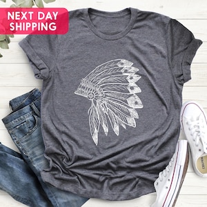 May include: A dark gray t-shirt with a white graphic of a Native American headdress. The t-shirt is laying on a white wooden surface with a pair of white canvas shoes.