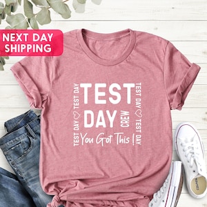 May include: A pink t-shirt with the text "TEST DAY CREW You Got This!" printed on it. The text is repeated on the sides of the shirt.
