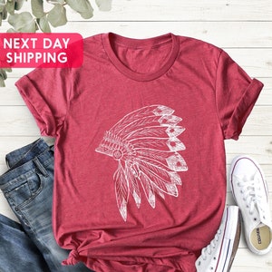 May include: A red t-shirt with a white graphic of a Native American headdress. The shirt is laying on a white wooden surface with a pair of blue jeans and white sneakers.