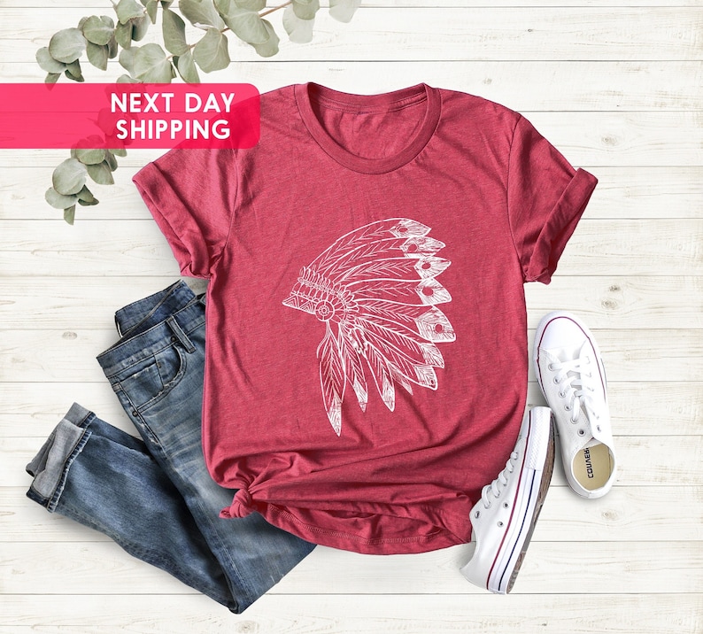May include: A red t-shirt with a white graphic of a Native American headdress. The shirt is laying on a white wooden surface with a pair of blue jeans and white sneakers.