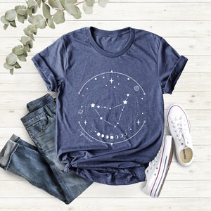 May include: A navy blue t-shirt featuring a white graphic of the Capricorn constellation, surrounded by a circle of stars and moons. The text "Capricorn" is written below the constellation.