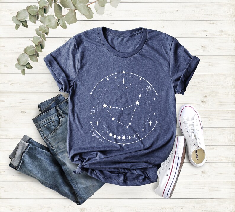 May include: A navy blue t-shirt featuring a white graphic of the Capricorn constellation, surrounded by a circle of stars and moons. The text "Capricorn" is written below the constellation.