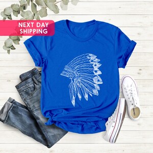 May include: A royal blue t-shirt with a white graphic of a Native American headdress. The t-shirt is laid flat on a white wooden surface with a pair of white sneakers and blue jeans.