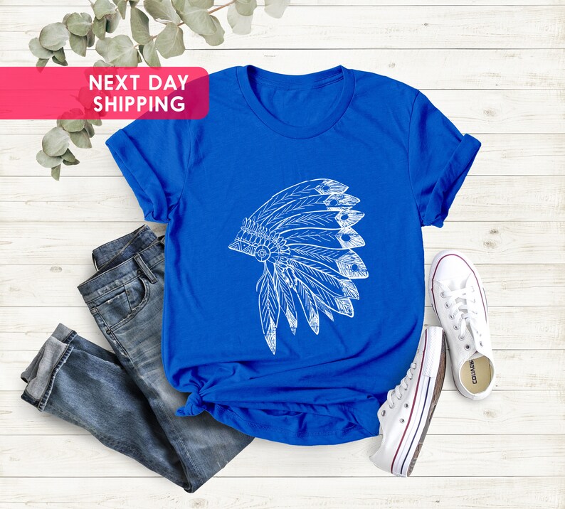 May include: A royal blue t-shirt with a white graphic of a Native American headdress. The t-shirt is laid flat on a white wooden surface with a pair of white sneakers and blue jeans.