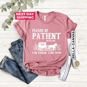 May include: A dusty rose t-shirt with a white graphic design that says "Please be patient with me I'm from the 1900s". The graphic features a vintage horse-drawn carriage with a horse and rider.