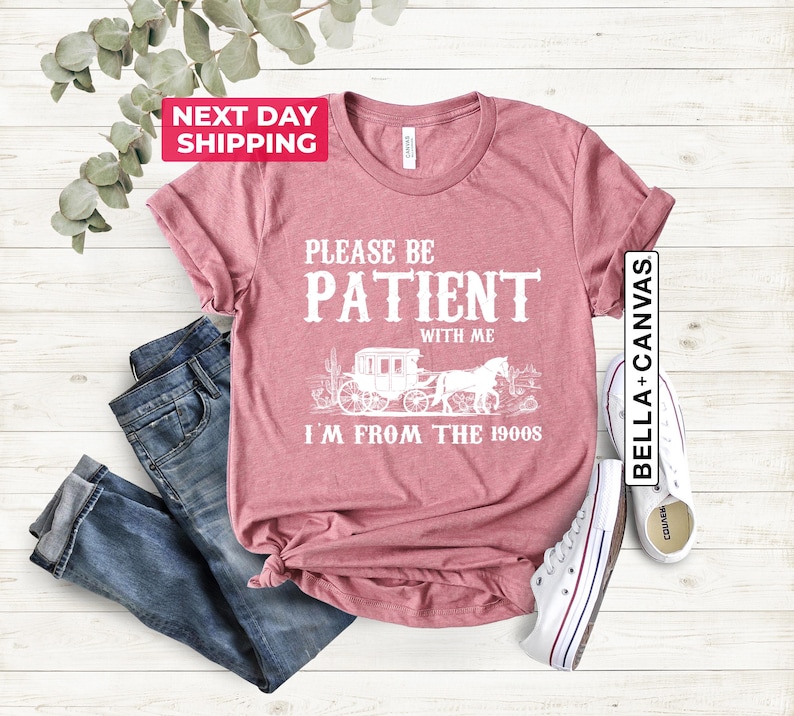 May include: A dusty rose t-shirt with a white graphic design that says "Please be patient with me I'm from the 1900s". The graphic features a vintage horse-drawn carriage with a horse and rider.