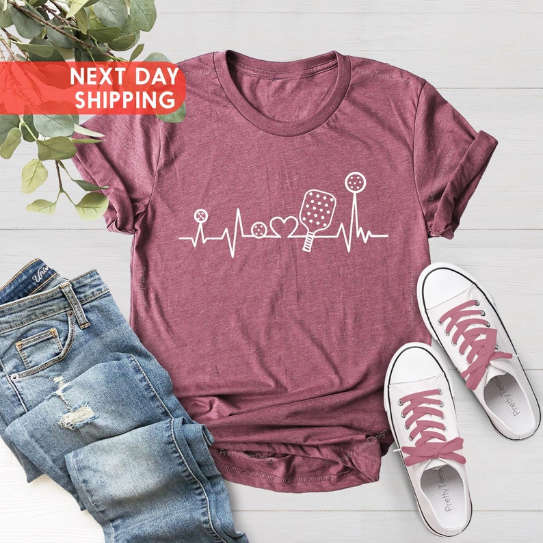 May include: A maroon t-shirt with a white graphic of a heartbeat with a pickleball paddle, ball, and heart in the middle. The text "NEXT DAY SHIPPING" is in a red box in the top left corner of the image.