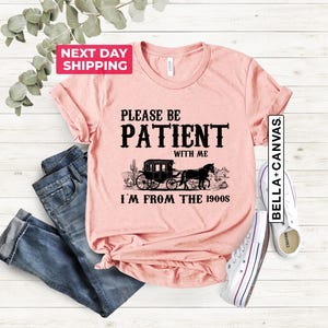 May include: A pink t-shirt with a black graphic that says "Please be patient with me I'm from the 1900s". The graphic also includes a black and white illustration of a horse-drawn carriage.
