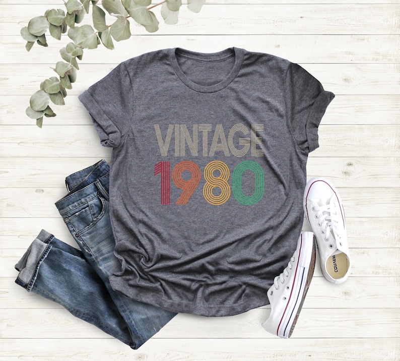 May include: A heather gray t-shirt with the text "VINTAGE 1980" in retro colors. The shirt is paired with blue jeans and white canvas sneakers. The design features a vintage aesthetic, perfect for a birthday or anniversary.