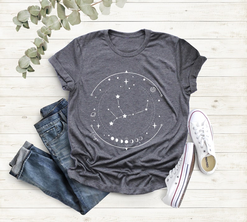 May include: A dark grey t-shirt featuring a white graphic of the Virgo constellation, encircled by a circle with stars, planets, and moons. The text "Virgo" is written beneath the constellation.