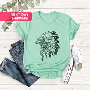 May include: A light green t-shirt with a black and white graphic of a Native American headdress. The t-shirt is laid out on a white wooden surface with a pair of blue jeans and white sneakers.