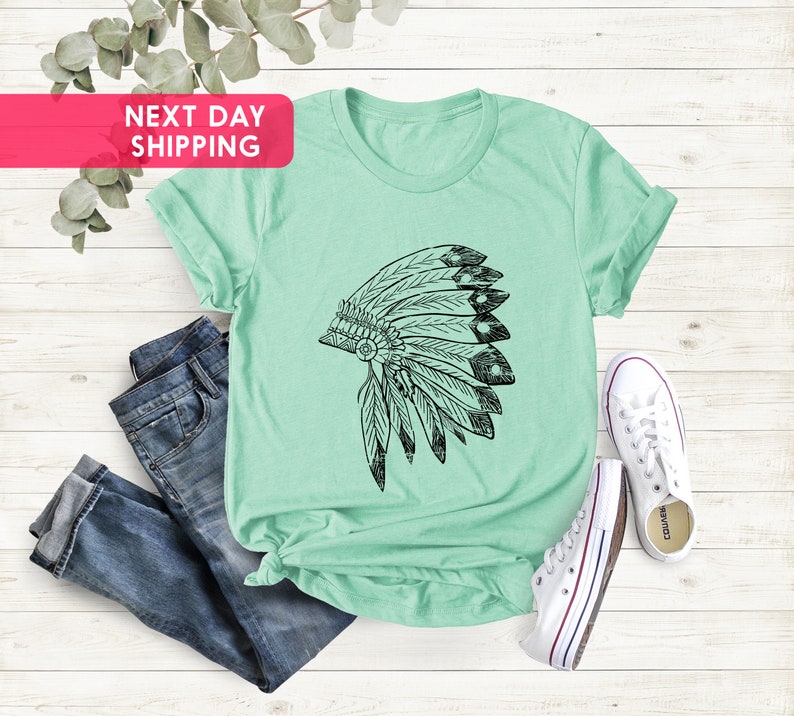 May include: A light green t-shirt with a black and white graphic of a Native American headdress. The t-shirt is laid out on a white wooden surface with a pair of blue jeans and white sneakers.