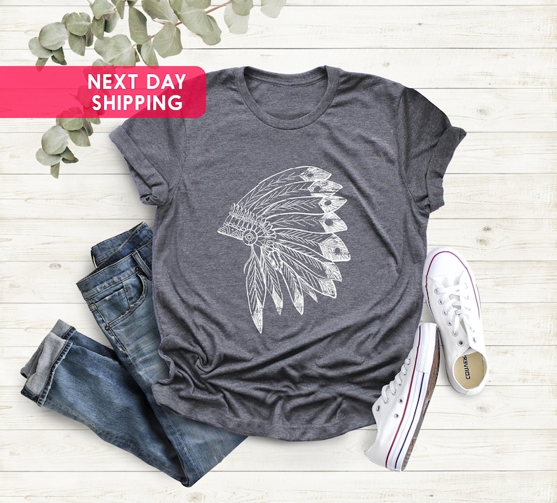 May include: A dark gray t-shirt with a white graphic of a Native American headdress. The t-shirt is laying on a white wooden surface with a pair of white canvas shoes.