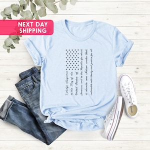 K&ouml;nnte beinhalten: Ein hellblaues T-Shirt mit einem schwarz-wei&szlig;en amerikanischen Flaggen-Design. Die Flagge ist mit dem Text des Treueids bedruckt.
