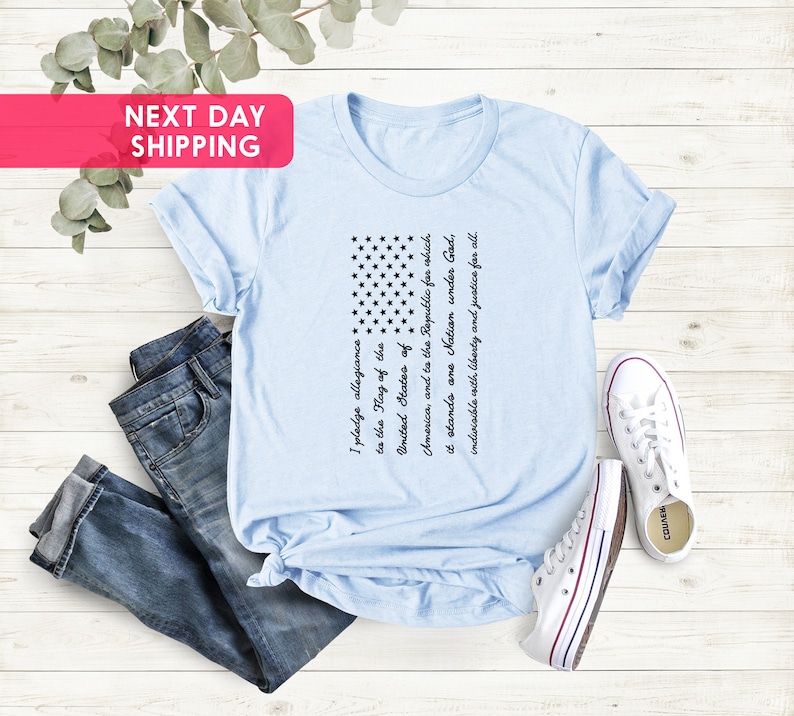 K&ouml;nnte beinhalten: Ein hellblaues T-Shirt mit einem schwarz-wei&szlig;en amerikanischen Flaggen-Design. Die Flagge ist mit dem Text des Treueids bedruckt.