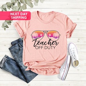 May include: Pink t-shirt with a graphic of sunglasses with palm trees reflected in the lenses. The text "Teacher Off Duty" is below the sunglasses.