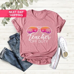 May include: A pink t-shirt with a white graphic of sunglasses with palm trees reflected in the lenses. The text "Teacher Off Duty" is below the sunglasses.