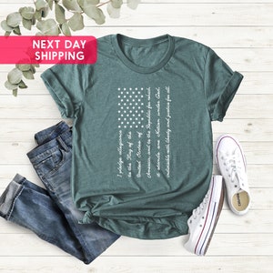 K&ouml;nnte beinhalten: Ein blaugr&uuml;nes T-Shirt mit einem wei&szlig;en amerikanischen Flaggen-Motiv und dem Text des Treueids darauf gedruckt. Das Shirt liegt flach auf einer Holzoberfl&auml;che mit einer blauen Jeans und wei&szlig;en Turnschuhen.