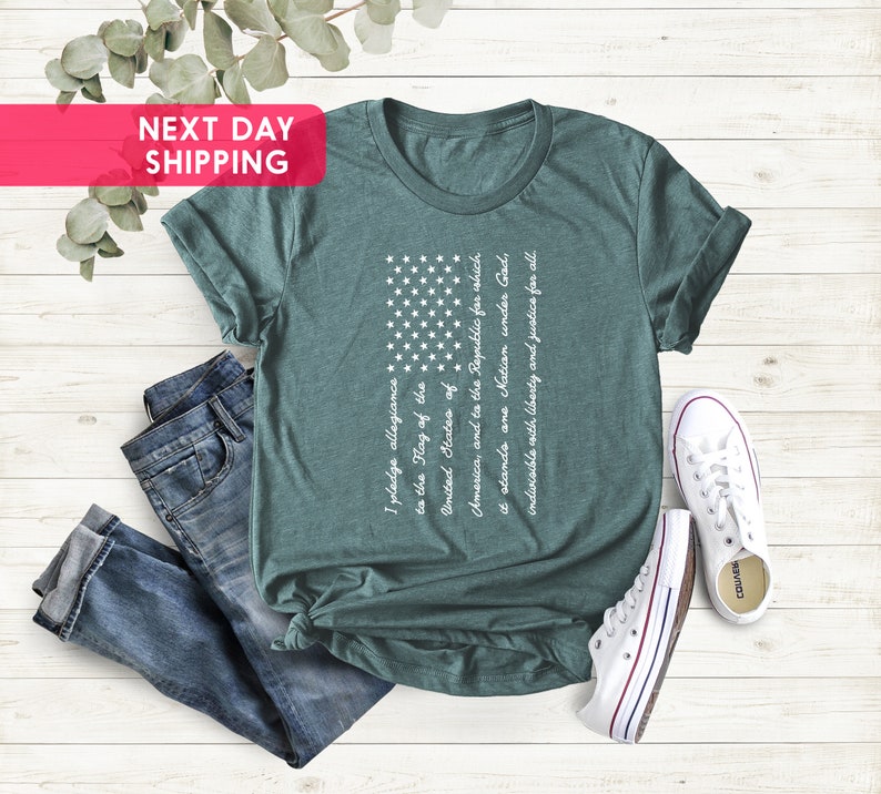 K&ouml;nnte beinhalten: Ein blaugr&uuml;nes T-Shirt mit einem wei&szlig;en amerikanischen Flaggen-Motiv und dem Text des Treueids darauf gedruckt. Das Shirt liegt flach auf einer Holzoberfl&auml;che mit einer blauen Jeans und wei&szlig;en Turnschuhen.