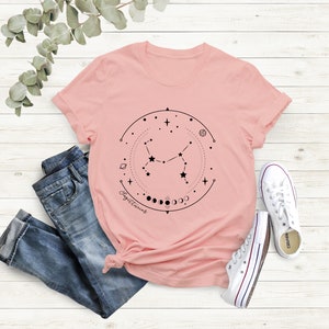 May include: A pink t-shirt featuring a black graphic of the Sagittarius constellation, moon phases, and the word "Sagittarius" printed on the front.