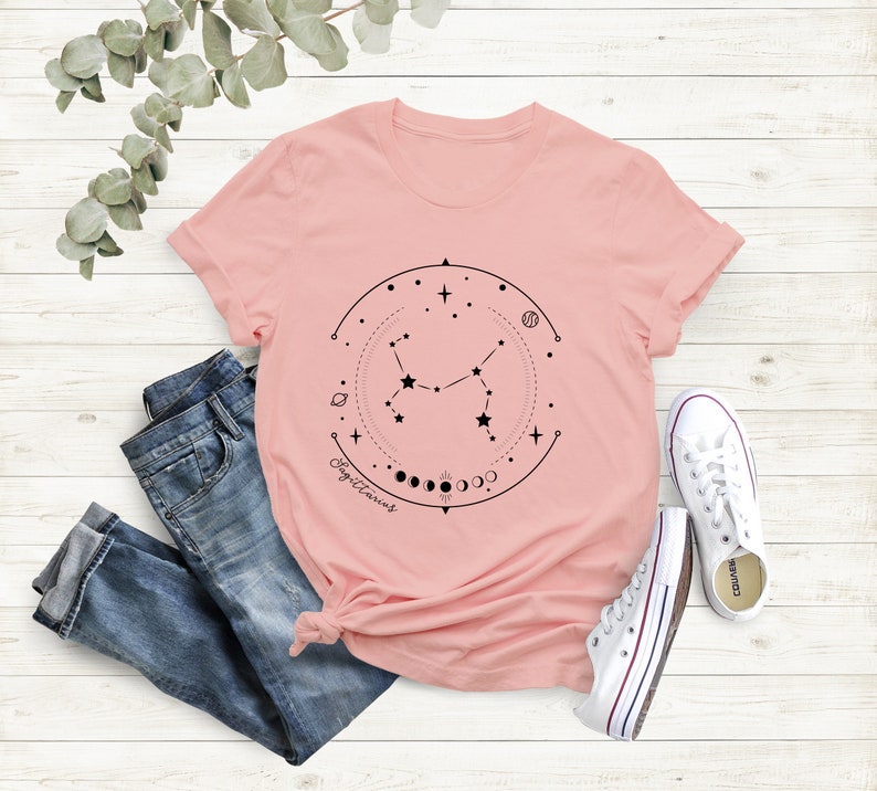 May include: A pink t-shirt featuring a black graphic of the Sagittarius constellation, moon phases, and the word "Sagittarius" printed on the front.