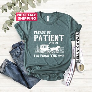 May include: Teal blue t-shirt with white text that reads "Please be patient with me I'm from the 1900s". The text is surrounded by a graphic of a horse-drawn carriage.