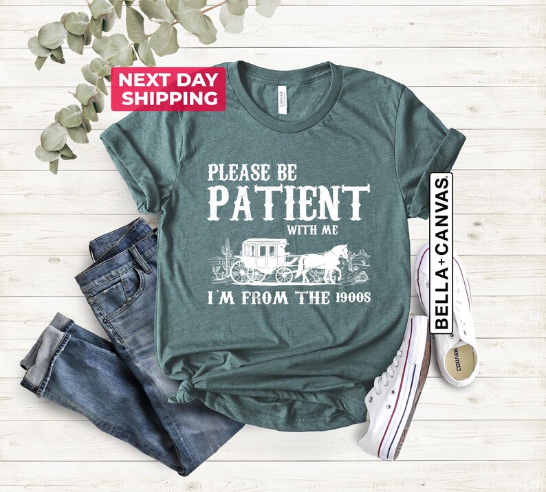 May include: Teal blue t-shirt with white text that reads "Please be patient with me I'm from the 1900s". The text is surrounded by a graphic of a horse-drawn carriage.