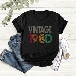 May include: Black t-shirt with the text "VINTAGE 1980" in a retro font. The numbers are in red, orange, and green. The shirt is paired with blue jeans and white sneakers.