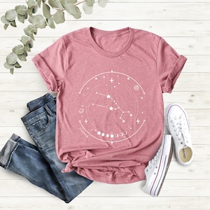 May include: A pink t-shirt featuring a white graphic design of a constellation with the text "Taurus" and a crescent moon phase.
