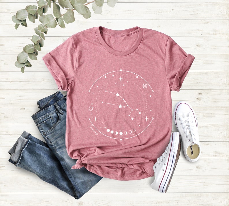 May include: A pink t-shirt featuring a white graphic design of a constellation with the text "Taurus" and a crescent moon phase.