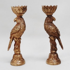 May include: A pair of gold-toned metal candle holders shaped like parrots. Each parrot is perched on a branch with a leaf-shaped candle holder on top.