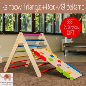 May include: A wooden rainbow triangle climber with a slide ramp and colorful rock climbing holds. The climber has a wooden handrail and is made of natural wood. The slide ramp is made of wood and has a colorful design. The text "BEST 1st Birthday GIFT" is on a red circle in the upper right corner of the image.