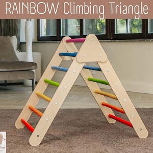 May include: A wooden climbing triangle with light-colored side panels and horizontal rungs painted in a rainbow gradient of red, orange, yellow, green, blue, and pink. The text RAINBOW Climbing Triangle is displayed at the top of the image.