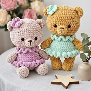 May include: Two handmade crochet teddy bears. One is light pink with a purple dress and flower. The other is tan with a mint green dress and flower. Both have black eyes and brown noses. A wooden star and vase with flowers are also in the image.