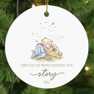 May include: A white circular ceramic ornament featuring a printed illustration of Winnie the Pooh and Eeyore hugging. The text reads: Thank you for making a difference in my story, followed by the year 2026. A gold string is attached for hanging.