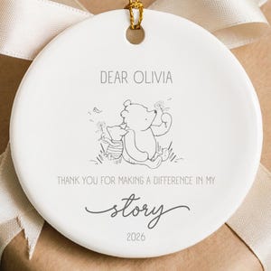 Puede incluir: Un adorno redondo de cerámica blanca con un dibujo lineal negro de Winnie the Pooh y Piglet. El texto dice: DEAR OLIVIA, THANK YOU FOR MAKING A DIFFERENCE IN MY story, 2026. Cuelga de un cordón dorado y está atado con una cinta blanca.