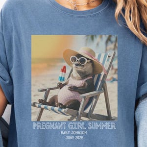 Puede incluir: Camiseta azul con un gráfico de una nutria con gafas de sol y sombrero, sentada en una silla de playa, sosteniendo un helado. El texto en la camiseta dice "Pregnant Girl Summer" con un nombre y una fecha.