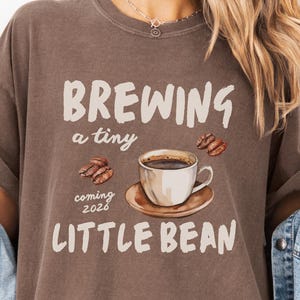 Może przedstawiać: Brązowa koszulka z napisem "BREWING a tiny LITTLE BEAN coming 2026" i grafiką filiżanki kawy. Koszulka ma luźny krój i podwinięte rękawy. Modelka ma na sobie dżinsową kurtkę.