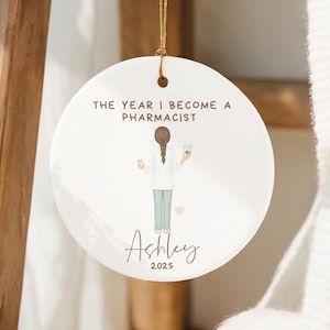 Puede incluir: Adorno redondo de cerámica blanca con una cuerda dorada, que representa a un farmacéutico. El texto "THE YEAR I BECOME A PHARMACIST" está encima de la ilustración, con "Ashley" y "2025" debajo. El adorno cuelga de una escalera de madera.