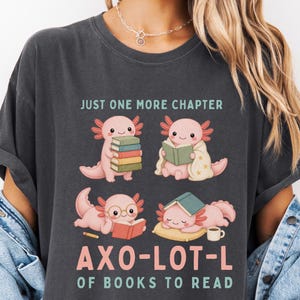 May include: A charcoal gray t-shirt featuring a graphic of four pink axolotls reading books. The text reads "JUST ONE MORE CHAPTER" and "AXO-LOT-L OF BOOKS TO READ" in pink and green lettering. The axolotls are shown holding books, wearing glasses, and resting.