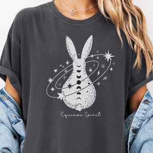 Puede incluir: Camiseta gris oscuro con un diseño blanco de un conejo rodeado de estrellas y fases lunares. El texto "Equinox Spirit" está debajo. La camiseta tiene un corte holgado y mangas enrolladas.