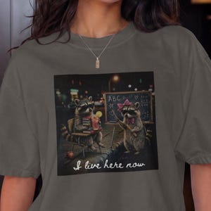 Op de afbeelding: Een antracietgrijs T-shirt met korte mouwen en een grafische print van twee wasberen, waarvan er één een drankje vasthoudt en de andere naar een schoolbord met de letters ABC wijst, inclusief de handgeschreven witte tekst I live here now.