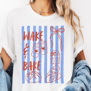 Puede incluir: Camiseta blanca con un gráfico rojo y azul. El gráfico presenta el texto "WAKE & BAKE" con ilustraciones de productos horneados y tazas de café. El diseño está sobre rayas verticales azules.