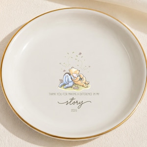 May include: A round white ceramic trinket dish with a gold-coloured rim. The centre features a small illustration of two cartoon bears sitting together, with the text "THANK YOU FOR MAKING A DIFFERENCE IN MY story" and the year "2024" printed below.