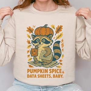 Könnte beinhalten: Beiges Sweatshirt mit einem Cartoon-Waschbären, der einen Kürbishut trägt und einen Taschenrechner hält. Der Text "PUMPKIN SPICE & DATA SHEETS, BABY." ist darunter gedruckt. Das Design enthält auch Herbstblätter und einen Kürbis.