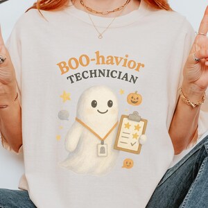 Könnte beinhalten: Cremefarbenes T-Shirt mit dem Text "Boo-havior Technician" in Orange und Gelb. Ein Cartoon-Geist mit Klemmbrett und eine Kürbis-Grafik sind ebenfalls abgebildet. Das Shirt ist ein Halloween-Design.